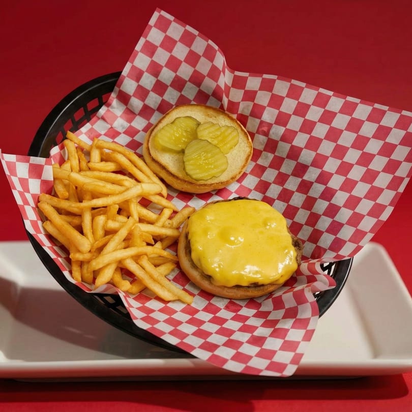 Cheeseburger with melted cheese, pickles, and a side of fries in a basket with red checkered paper.